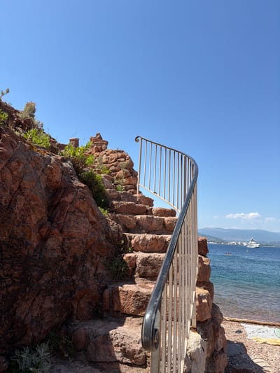 The image shows a curved staircase made of natural red stones leading up a rocky incline. The staircase is bordered by a metal railing with vertical bars. Green vegetation grows in patches between the rocks and on the staircase. To the right of the stairs, a clear blue sky stretches above the turquoise sea. A large white yacht is visible on the water in the distance, along with a coastline dotted with buildings. A small beach with pebbles and some beach towels is visible in the foreground on the right. The overall scene is a picturesque coastal view in Théoule-sur-Mer, France, likely taken on a sunny day.