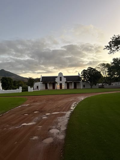 The image displays a picturesque landscape in Drakenstein Nu, South Africa, featuring a classic Cape Dutch-style building. The prominent structure is a white, multi-gabled building with dark roofs and traditional dark wooden shutters or doors, set against a backdrop of mountains. Several large, dark green trees, some appearing ancient, are scattered around the property, partially obscuring the building.

In the foreground, a wide, reddish-brown dirt path, characteristic of a country estate or farm road, stretches into the distance. This path is notably wet and muddy, with numerous puddles reflecting the cloudy sky, indicating recent rain. Alongside the dirt path, a well-maintained, lush green lawn runs parallel to the building.

The sky is dominated by large, dramatic clouds, with a soft, warm glow visible towards the horizon, suggesting either dawn or dusk. The light illuminates the edges of the clouds and casts a gentle glow over the scene. Mountains rise in the distant background, their contours visible beneath the cloud cover. There are no people visible, and no specific activity is taking place, creating a serene and timeless atmosphere. No visible text is present in the image.