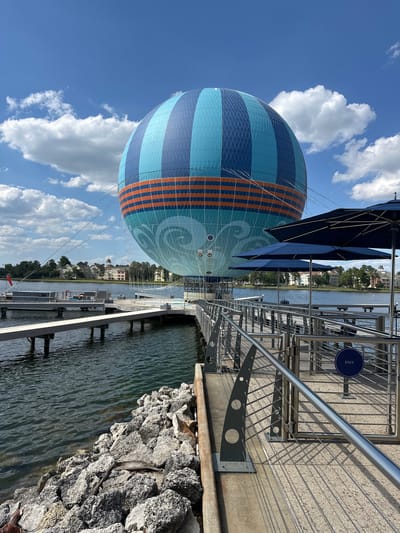 The image is a landscape photograph taken in Lake Buena Vista, Florida, United States. In the foreground is a partially visible, inflated, striped, blue and orange hot air balloon, anchored to the ground. Beside the balloon is a metal barrier, a row of dark-blue umbrellas, and a stone wall with the water of a lake or river on the right. The background includes trees, buildings, and a mostly clear blue sky with some white clouds. The composition includes both natural elements like the sky and water and man-made structures like the balloon and the barriers. The photo is taken from a relatively low angle, emphasizing the size of the balloon.