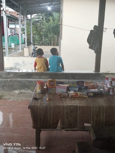 Two young boys are sitting on a concrete ledge outside a home in Anjatan, Indonesia (latitude: -6.419, longitude: 107.978). The boys are facing away from the camera, and the one on the left is wearing a yellow shirt, while the one on the right is wearing a blue shirt. They are looking at something off-camera, and there is a table in front of them. The table is covered with a brown tablecloth and is laden with various packaged snacks and drinks. The scene is set in a partially covered outdoor area with a view of a backyard and a partly obscured bicycle. The floor is a mixture of concrete and tiles. The atmosphere is relaxed and casual.  A single brown jacket is hanging on a post in the background.  The image was taken on March 14, 2023, at 15:47.
