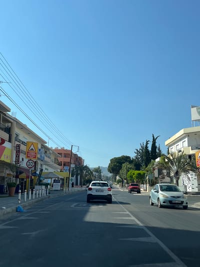 The image depicts a street scene in Germasogeia Municipality, Cyprus, likely during the daytime under a clear blue sky, suggesting fair weather. The perspective is from within a vehicle, looking forward and slightly to the right.
The scene is characterized by buildings lining one side of the street, with a few cars visible. On the left side of the road, there are multi-story buildings with shops and businesses on the ground floor. Signs with text are visible on these buildings, including some with Greek lettering, such as "ΚΥΠΡΙΑΚΗ ΟΔΟΣ," which translates to "Cypriot Road," and a sign indicating "Road Humps." A yellow triangular sign with a black border and an image depicting road humps is also prominent. Further down the street, another circular sign with a red border and the number "30" suggests a speed limit.
On the right side of the road, a light blue car with the license plate "MHZ 175" is moving away from the camera. Behind it, a dark-colored car and a red vehicle are also in motion. Further down, a white car with the license plate "TEH 292" is parked. The road itself has lane markings and a shadow of a vehicle is cast across it.
Trees, including palm trees and other greenery, are interspersed between the buildings and along the roadside, adding a touch of nature to the urban environment. The presence of power lines strung across the scene indicates urban infrastructure.
The overall impression is a typical street scene in a Mediterranean town, with commercial activity and traffic. There are no individuals clearly visible or identifiable as the primary subject. The time of day appears to be morning or afternoon, judging by the quality of the light and the shadows.