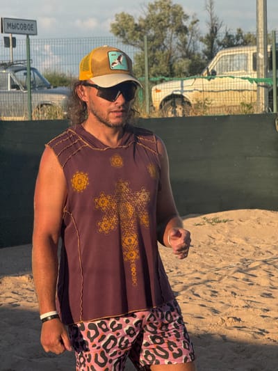 A medium shot captures a man standing on a sandy beach. He is wearing a sleeveless hooded t-shirt with a geometric pattern and leopard-print shorts. A baseball cap with a patch is on his head, and he has sunglasses on. The man is looking to his right, with his left arm bent at the elbow and his hand outstretched. In the background, a fence separates the beach from a street with a sign that reads "МЫСОВОЕ," and two parked cars. Trees and a white vehicle are visible behind the fence. The sun is casting long shadows on the sand.
