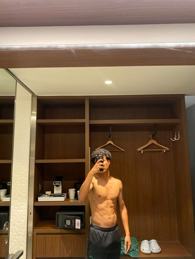 A shirtless, muscular man stands in a wooden hotel closet, taking a photo of himself with a phone. The closet contains two hangers, a coffee maker, a safe, a folded green towel, and a pair of white clogs. A large mirror reflects the lower portion of the closet and the floor. The closet door is a light color with brown trim. A light fixture is visible on the wall to the left of the closet.