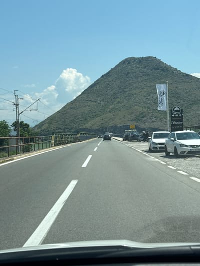 This media file captures a roadside scene in Kruševica, Montenegro, on a clear, sunny day. The perspective is from inside a vehicle, looking out at a road that runs alongside a steep, green mountainside.
On the left side of the road, a white flag with a black emblem is prominently displayed, possibly indicating a business or event. Further along, a sign for "RESTORAN STARČEVO" with a fish emblem is visible, suggesting a restaurant. A few vehicles are parked along the roadside, including two white cars and a motorcycle with a rider. The road itself is paved with asphalt and marked with white lines. A metallic fence runs along the edge of the road, separating it from the embankment. In the distance, a power line structure with wires is visible against the blue sky, which is dotted with a few fluffy white clouds. The overall impression is of a bright, warm day, likely in the afternoon, given the lighting.
