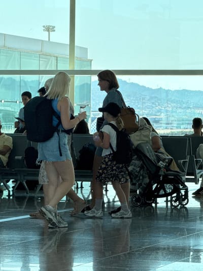 The image shows several people sitting and standing in what appears to be an airport terminal. A stroller is visible, suggesting travel with a child or for convenience. The setting is indoors, with large windows looking out onto a landscape with buildings and hills, possibly indicating a view of the city and its surroundings. The lighting suggests it is daytime. The people are dressed casually, some with backpacks and bags, indicating they are travelers. One person is wearing a blue top and denim shorts, another a floral print skirt, and another a dark hoodie. The floor is reflective, creating mirrored images of the people and objects. The overall mood seems relaxed, as people are waiting. There is no visible text in the image. The location context provided is el Prat de Llobregat, Spain, which is known for being the location of Barcelona–El Prat Airport.
