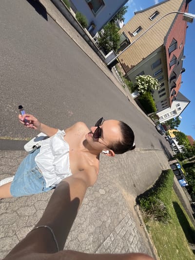 The image shows a woman taking a selfie on a street in Saarbrücken, Germany. She has dark hair pulled back, is wearing sunglasses and a white off-the-shoulder top with a denim skirt. She is holding what appears to be an electronic cigarette. In the background are residential buildings, trees and parked cars. The street is paved with asphalt and the sidewalk is paved with interlocking stones. The weather is sunny.