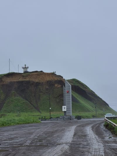 The image captures a view of a memorial and a lighthouse situated on a grassy hillside overlooking a wet, unpaved road. In the foreground, the road is muddy and has several puddles, indicating recent rain. A white car with an open driver's side door is parked on the right side of the road, facing away from the camera, near a metal guardrail. Further back, a concrete and stone monument with a red star is visible. To the left of the monument, a lighthouse with a white tower and a red-capped lantern room stands on the crest of the hill. The hillside is covered in green grass, with visible erosion marks on the steeper sections. The sky is overcast and a light gray, suggesting a cloudy or foggy day. The scene appears to be a desolate, coastal area. This location is identified as being near the northern end of Uglegorsk, Russia, likely a historical site given the presence of the memorial and lighthouse.