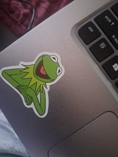 An overhead, angled view captures the upper surface of a light grey metallic HP EliteBook laptop. A sticker of Kermit the Frog, depicted smiling with both hands clasped beneath his chin, is affixed to the laptop's left palm rest area. The sticker features a green frog, a white eye outline, and a pink mouth with a red tongue, all outlined in white. To the right, a portion of the laptop's black keyboard is visible, showing keys labeled "ctrl," "shift ↑," "fn lock," and the Windows logo. The lower right segment of the laptop's trackpad area is also visible. The laptop rests on a textured light grey fabric, possibly clothing or upholstery. In the blurred background, a dark maroon fabric object and a black electrical cable are discernible. The scene is located in Kahawa Sukari ward, Kenya.