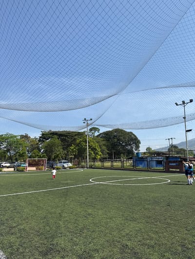 The image displays an outdoor synthetic turf soccer field located in Pitalito, Colombia, under a clear blue sky. A large black netting is suspended overhead, covering the playing area. The field is marked with white lines, including a center circle and segments of penalty boxes. On the left side of the field, a small soccer goal with a red frame and white net is visible. One small child, dressed in a white shirt and red shorts, walks on the green turf, positioned near a soccer ball. On the right side of the field, two individuals stand near the sideline; one wears a light blue jersey, white shorts, and black socks. The background features dense green foliage and trees. Behind a fence on the left, multiple vehicles are parked, including a white SUV and a blue car. In the mid-ground right, beyond a black metal fence, a blue building with a reddish-brown roof is discernible. Tall poles equipped with floodlights are present on both sides of the field, and distant hills or mountains can be seen on the horizon.