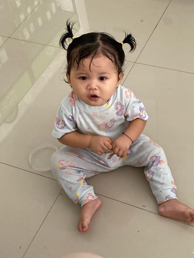A young child with dark hair styled in two pigtails is sitting on a tiled floor, which appears to be in an indoor setting. The child is wearing light blue pajamas adorned with cartoon characters. The child's facial expression is neutral, and they are looking directly at the camera. Their hands are clasped together in front of them. The floor is composed of large, light-colored tiles, with visible grout lines. A translucent plastic ring is visible on the floor to the left of the child. The background shows some reflective surfaces, possibly a wall or furniture, with subtle light and shadow patterns. The image was taken in Ta', Indonesia.