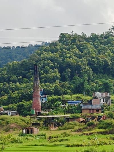 The image depicts a rural landscape in Tharpu, Nepal, characterized by lush green hills and vegetation. In the foreground, a field of bright green crops, likely rice paddies, stretches across the lower portion of the frame. A prominent feature of the scene is a tall, red brick chimney, typical of a kiln, rising from a cluster of buildings on the side of a hill. The chimney tapers towards the top and appears aged, with some staining evident.
Scattered around the base of the chimney and on the hillside are several structures. To the right of the chimney, a multi-story building with a white facade and multiple windows is visible. Adjacent to it, a smaller brick building with a blue roof is partially obscured by foliage. Further down the slope, various other low-lying structures, some appearing unfinished or industrial in nature, are present. Construction equipment, including a yellow backhoe loader and another piece of machinery, are situated amongst the buildings and disturbed earth, suggesting ongoing construction or excavation. A green water tank is mounted on a support structure near the chimney. Power lines crisscross the upper part of the image, running horizontally above the treeline. The background is dominated by a dense, green forested hillside that rises steeply. The sky is overcast and bright, suggesting diffused daylight. No people are visible in the image.
