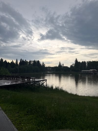 A cloudy, overcast day is depicted at a lake, possibly in or near Chusovoy, Russia. The foreground features a wooden pier with several empty lounge chairs, suggesting a recreational area. The pier is wet, indicating recent rain. Beyond the pier, the lake's surface is rippled, with small circles visible, confirming it is raining. A dense line of trees forms the treeline on the far side of the lake, obscuring any distant structures except for a building with a distinct architectural style, possibly a church with two spires, visible further in the distance. To the right, a floating structure, perhaps a boathouse or restaurant, is situated on the water. The overall atmosphere is calm and muted, typical of a rainy or pre-rainy day in a natural setting.
