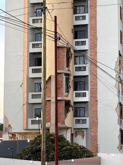 The image shows a tall building in Maracay, Venezuela, with visible structural damage. Large cracks run down the exterior walls, and sections of the facade have crumbled, exposing the brickwork and concrete structure beneath. The damage appears to be concentrated on one side of the building, suggesting a localized event or structural failure.
Several windows, some with protective grates, are visible on the undamaged portions of the building. Air conditioning units are installed in some of the window openings. A telephone pole with multiple wires is situated in front of the building, obscuring a portion of the damaged facade. Behind the pole, a dense cluster of flowering bushes with pink blossoms is visible, along with a section of a brick wall and some electrical fencing. The sky is overcast, and the overall lighting suggests it is daytime. There are no people visible in the image.