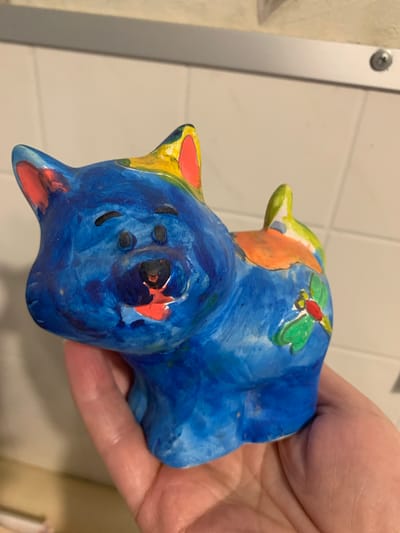 The image shows a close-up of a brightly painted ceramic figurine of a dog. The dog is primarily blue with accents of yellow, orange, and green. It has distinct red inner ears and a red tongue. There is a small green and red insect-like design on its side. The figurine is being held in a person's hand, which is visible in the lower portion of the frame. The background appears to be a tiled wall, suggesting an indoor setting, possibly a home or a craft studio. There is no visible information in the image to indicate its location in Відзаўскі сельскі Савет, Belarus.