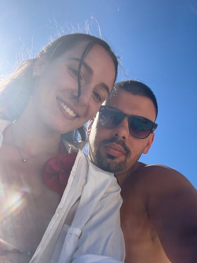 A selfie taken from a low angle featuring a man and a woman against a clear blue sky. The woman is in the foreground, smiling and looking at the camera. The man is behind her, wearing sunglasses and with a visible beard. He is shirtless, and the woman is wearing a white shirt over a red top, with a silver necklace. The location appears to be outdoors, potentially near a beach or coastline in el Poble Nou de Benitatxell / Benitachell, Spain. The bright sunlight indicates it is likely daytime.