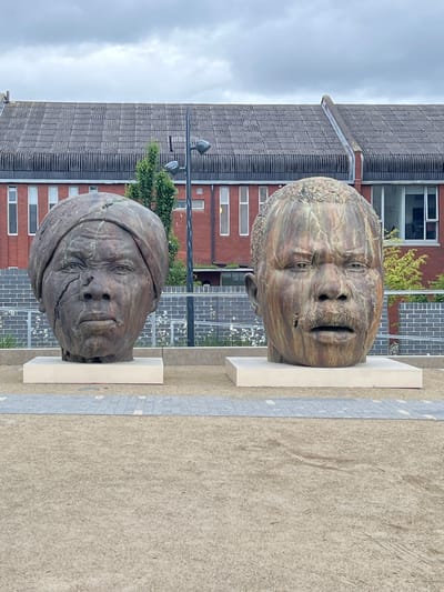 The image captures two oversized, bronze head sculptures displayed on white plinths in an outdoor urban setting. These sculptures appear to depict human faces. The top head has a textured surface reminiscent of wood grain. The lower head appears wrapped with a scarf or a similar covering.
Behind the sculptures, there is a modern building featuring red brick and multiple windows. A streetlight is also visible in the background. The ground is paved with light tan-colored stones.
Without further information, pinpointing the exact location within Dublin cannot be accomplished.