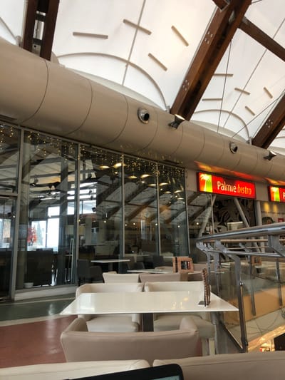 The image, originally rotated 90 degrees clockwise, depicts the interior of a modern commercial space, likely a bistro or dining area within a larger building such as a shopping mall in Marousi, Greece. A prominent, tall, cylindrical column, light beige in color, runs through the center of the frame, adorned with spotlights and circular vents.
Above and to the left of the column, a high ceiling features a striking design with white fabric panels forming circular and linear patterns, supported by sturdy dark wooden beams. To the right, a multi-level glass partition or wall provides a view into an adjacent area, possibly an escalator well or another floor. This glass wall is illuminated by decorative string lights, casting a soft glow. Through the glass, dark tables and chairs are visible, along with a distant window offering a glimpse of the exterior, including a plant with red leaves.
In the immediate foreground on the right, a dining area is furnished with light-colored, possibly cream or beige, upholstered chairs and sleek white tables. A metal handrail borders this seating area. A black screen or tablet is partially visible on the far right edge, potentially displaying information or acting as an e-reader.
A noticeable red illuminated sign on the column clearly displays "Palmie bistro" in white text, accompanied by a palm tree logo. Inside the glass-enclosed area, another rectangular display shows the text "TV+YOU" and partially visible words like "Green history and pro...". The scene is well-lit, suggesting daytime or a brightly illuminated interior during evening hours. No people are present, indicating a tranquil moment in the establishment.