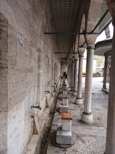 The image shows a row of stone fountains and benches located under a covered walkway in the courtyard of the Süleymaniye Mosque in Istanbul, Türkiye (latitude: 41.027, longitude: 28.98). The mosque is a 16th-century Ottoman imperial mosque commissioned by Suleiman the Magnificent. The image shows a row of stone fountains with metal taps and a series of stone benches. The fountains are attached to a long wall, while the benches are positioned in the middle of the courtyard, aligned with the row of stone pillars that form the covered walkway. The walkway is made of stone with a tiled ceiling. The scene is quiet and the only person visible is in the distance, walking away from the camera. The image is captured from a low angle looking along the row of fountains towards the covered walkway.