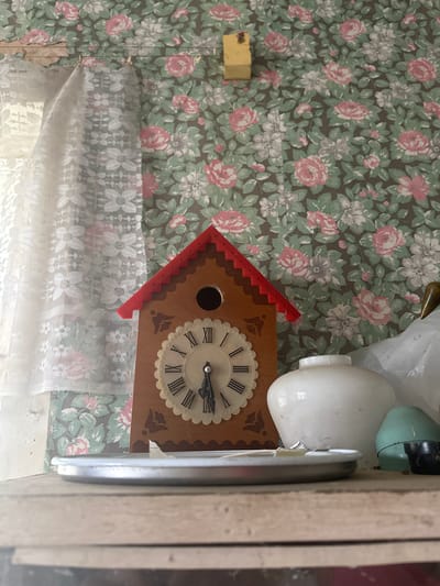 The image shows a cuckoo clock hanging on a wall with floral wallpaper. A piece of yellow foam is attached to the wallpaper. A white lace curtain hangs below the clock. Other objects, including a white light bulb, are partially visible to the right of the clock. A light-colored wall is visible in the far right corner of the image. The scene is indoors.