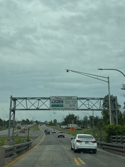 In Town of Grand Island, United States of America, on a cloudy day, a car is driving on a multi-lane highway under a toll plaza overhead sign. The sign indicates "TOLL - CARS" and lists prices for E-ZPass and Tolls by Mail. The E-ZPass price is $1.00 for New York and $1.75 for Other. Tolls by Mail is $1.75. There is also a notice for a "HIGHER TOLL FOR ADDITIONAL AXLES." Several cars are visible on the highway, moving in the same direction. The environment consists of a paved road, guardrails, trees, and utility poles. The sky is overcast with thick clouds.