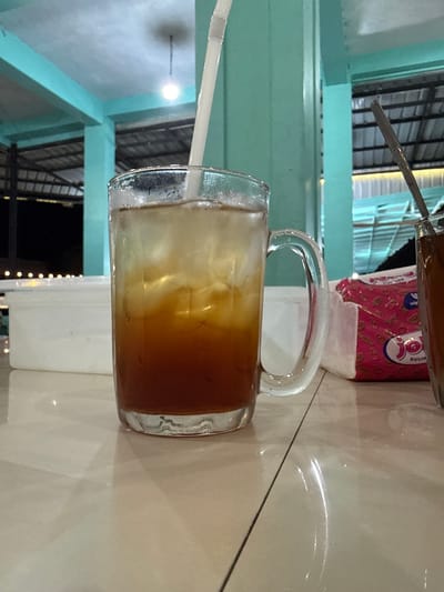 A glass of iced tea sits on a tiled surface in front of a white container. The drink, with ice cubes and a straw, is dark brown at the bottom and lighter at the top, suggesting it is iced tea. To the right, another glass contains a dark liquid, likely coffee or tea. A pink and white tissue packet with a logo is visible between the glasses. The background features turquoise pillars and a roof structure, suggesting an outdoor or semi-outdoor setting, possibly a food court or restaurant in Grong-Grong, Indonesia. The lighting is dim, with a single bulb visible in the distance, and the overall atmosphere is casual.