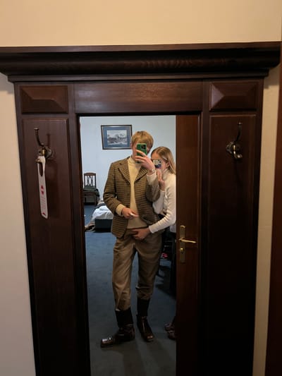 A couple poses in a mirror selfie. The man, with blonde hair, wears a tan turtleneck, plaid blazer, tan pants, and riding boots. He holds a smartphone with a green case. The woman, with blonde hair, wears a white top and holds a smartphone. The reflection shows a hotel room interior with blue carpet, a framed picture, and various furniture. The mirror is part of a dark wood structure with hooks and a door handle.
