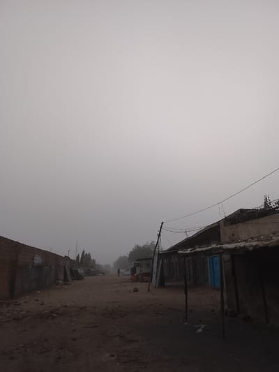 A desolate dirt road stretches into a dense fog, flanked by worn buildings. A lone figure walks in the distance, barely visible through the heavy haze. To the left, a brick wall stands alongside debris. On the right, dilapidated structures with decaying canopies line the street. Power lines sag above the scene, contributing to the atmosphere of decay and isolation. The overall impression is one of neglect and stillness, obscured by the pervasive fog.