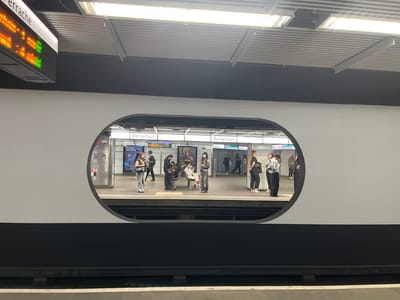 The image shows a view of the Bellecour metro station platform in Lyon, France, as seen through an oval-shaped window on the side of a metro train. Several people are standing or sitting on benches on the platform, waiting for a train. Some are looking at their phones, while others are talking to each other. The station has signage that says "Bellecour". The architecture is modern, with white walls and a black lower section. The platform is well-lit. A digital display sign indicating the metro station "Perrache" is visible on the top left of the image.