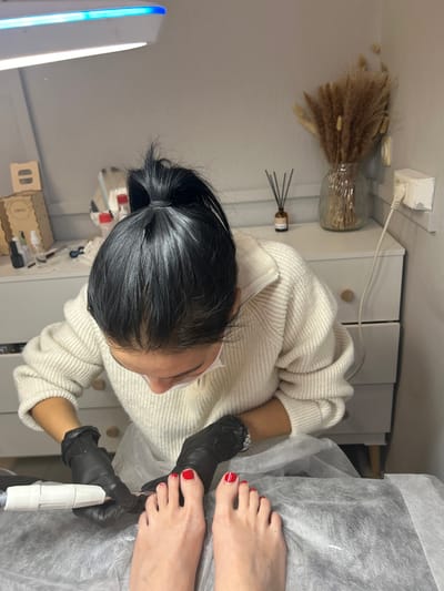The image portrays an indoor scene within a beauty salon located in the city of Voskresensk, Russia. A pedicurist, wearing a light-colored ribbed sweater, a white face mask, and black gloves, with dark hair tied in a high ponytail, is depicted leaning over to perform a pedicure. The pedicurist's right hand grips a white electric nail file/drill, positioned towards the client's left foot. The client's bare feet are visible in the foreground, resting on a disposable grey protective sheet. All ten toenails are polished red, with the exception of the client's right big toe, which features a white stripe over the red polish. The background includes an overhead white and blue LED lamp illuminating the workspace. To the left, a wooden box, small bottles, and salon tools are arranged on a surface. To the right, a white dresser displays an amber-colored reed diffuser bottle and a clear glass vase filled with dried pampas grass, with a white electrical outlet and cord visible on the wall. The walls are light grey.