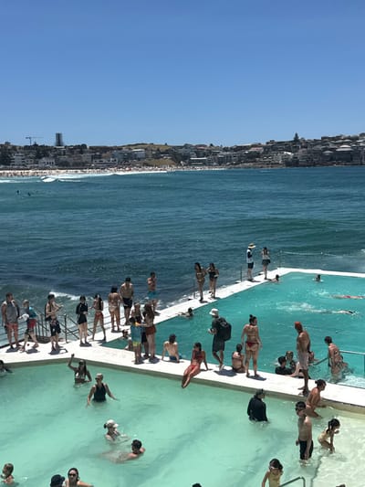 A coastal scene features an ocean pool teeming with people enjoying the water and sun. The pool's edge is lined with individuals sunbathing and relaxing. Beyond the pool, the ocean stretches towards a shoreline dotted with buildings. A clear blue sky dominates the upper portion of the image, creating a vibrant and lively atmosphere. The composition captures a moment of leisure and recreation at a popular seaside destination.