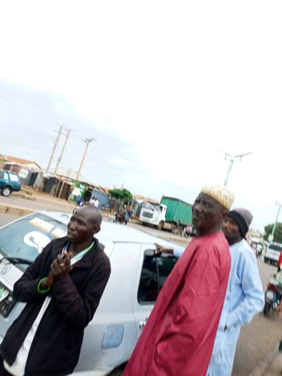In Tumfure, Nigeria, a group of men are gathered near a street. One man in the foreground wears a black jacket over a white shirt and is holding his hands clasped. To his right, another man in a maroon robe and a patterned cap faces away from the camera. Behind him, a third man in a light blue outfit stands with his hand in his pocket. A silver car is parked on the left side of the image. In the background, there are buildings with corrugated metal roofs, power lines, and several trucks. The sky is overcast.