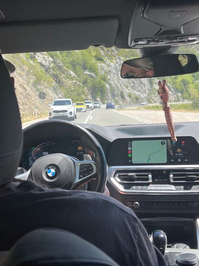 The image captures the interior of a BMW, looking forward from the driver's perspective. The driver, wearing a black t-shirt, is partially visible in the foreground. The car is traveling on a road with a steep, rocky, and vegetated mountainside on the left and a similar landscape on the right. Several cars are visible ahead on the road, including a white SUV, a yellow car, and a blue sedan. The car's dashboard displays navigation on a central screen, showing Google Maps and the current speed as 98 km/h. The driver's instrument cluster shows a speed of 100. A decorative, pink, doll-like object hangs from the rearview mirror. The scene suggests the car is driving on a scenic route, possibly through a mountainous region, in Brajici, Montenegro.