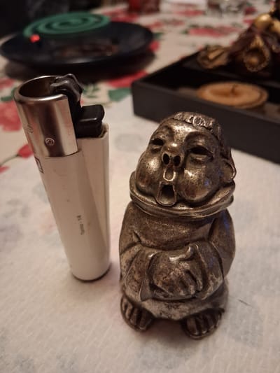 A close-up shot shows a metal figurine of a gargoyle lying on a patterned tablecloth. The figurine is detailed with a grotesque face, emphasizing its features like eyes, nose, and mouth. Behind the figurine, a black rectangular object is visible, possibly a box, with two round, light-colored objects inside, resembling coasters or small decorative plates.
In the foreground, a white and silver disposable lighter rests on the tablecloth. The lighter's metallic base is clearly visible, and the white plastic body has some text imprinted on it. The text on the lighter reads "Torino - 1/4". To the left of the lighter, a dark circular object with a green spiral pattern is partially visible, with a small red light emitting from its center. This object might be an electric mosquito repellent device or a similar item.
The lighting in the image suggests it was taken indoors, possibly in the evening, as there is no indication of natural daylight. The overall scene appears to be a still life arrangement of decorative items. The setting, with the patterned tablecloth and the objects, gives a sense of a domestic or personal space. The figurine's expression could be interpreted as menacing or humorous, depending on the viewer's perspective. No specific activity is taking place; the objects are simply arranged.