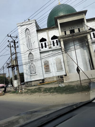 This media file captures a partially constructed building, possibly a mosque, with a prominent green dome. The structure features white ornate windows and intricate white latticework. In the foreground, a road is visible, and the image appears to be taken from a vehicle, as indicated by the reflection on the car's surface. Utility poles and numerous electrical wires crisscross the frame, suggesting an urban or semi-urban setting.

To the left of the building, a small stall with a pink and white umbrella is set up, and a person is seated nearby. Behind the stall, a white car is parked. The ground is a mix of dirt and grass, with some construction materials visible. The sky is overcast, indicating a cloudy day. The overall scene suggests a daytime setting, likely during business hours given the presence of the stall and parked car. No visible text is discernible in the image. The architectural style and the presence of the mosque-like structure, along with the general environment, hint at a location in a region where Islam is prevalent, consistent with the provided location context of Keudelapang, Indonesia.