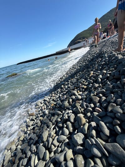 The image shows a sunny day at a pebble beach in Partenit, Ukraine. The foreground is dominated by a dense field of smooth, grey pebbles, some of which are wet from the lapping waves. The sea to the left is a muted blue-green, with white foam washing onto the shore. In the background, people are visible on the beach and in the water. A woman in a colorful bikini walks down the pebble slope towards the sea. Further back, a group of people are seen on the beach, some sitting and some walking. A long, dark breakwater extends into the sea on the left side of the frame. A steep, tree-covered hill rises sharply behind the beach on the right. The sky is a clear, bright blue.
