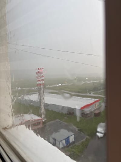 The image is a blurry, overcast outdoor scene viewed through a wet window. In the middle ground, a tall, red and white striped tower, possibly a communication tower, stands prominently. To the right of the tower is a long, low-rise building with a red roof and some green accents, appearing to be a commercial or industrial structure. Further to the right, a silver vehicle is parked. In the foreground, water streaks on the window obscure the view. The background consists of a hazy, indistinct landscape with what appear to be roads and distant lights. The overall impression is one of a dreary, possibly rainy day in an industrial or semi-urban area. This scene is located in Засечное, Russia.
