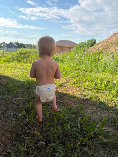 A young child, seen from behind, stands barefoot in a grassy area. The child is wearing a diaper and has blond hair. The background shows a wooden structure, likely a shed or outbuilding, to the left. Beyond that, a blue sky with white clouds is visible, along with power lines and distant trees. The scene appears to be set outdoors in a rural or semi-rural location, possibly a backyard or field, given the presence of grass, dirt, and vegetation. The lighting suggests it is daytime, with sunlight casting shadows. There is no visible text in the image. The child's posture and the outdoor setting suggest a moment of play or exploration.