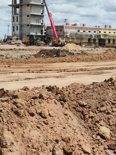 This media file captures a construction site in Arkalyk, Kazakhstan, under a cloudy sky, likely during daylight hours. The scene is dominated by the ongoing construction of buildings and the surrounding earthwork.

In the background, a multi-story concrete structure stands incomplete, with exposed rebar and scaffolding. Adjacent to it, another building with brickwork and yellow insulation is under construction, also featuring scaffolding. A prominent red crane is positioned near the concrete structure, its boom extended upwards. Several construction vehicles are visible, including excavators and a wheel loader. A person in dark clothing is walking near the wheel loader.

The foreground and middle ground are filled with piles of reddish-brown soil, indicating significant excavation and earthmoving activities. Tracks from heavy machinery are clearly visible on the cleared dirt surfaces, suggesting the movement of equipment and materials.

The weather appears overcast, with a mix of clouds in the sky, but the lighting suggests it is daytime. The overall impression is one of active construction, with raw materials and heavy machinery defining the landscape. No visible text is present in the image.