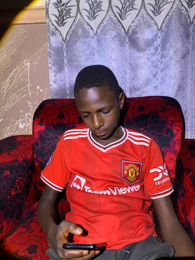 A young boy is sitting on a red patterned armchair, holding a black smartphone in his left hand. He is wearing a red Manchester United jersey with white trim on the collar and sleeves. The jersey has the Manchester United crest on the left chest and the TeamViewer logo across the front. The wall behind him is decorated with a grey patterned curtain and a section of wall with white paint splatters. The lighting suggests an indoor setting, possibly a home in Barakin Sabawa, Nigeria.
