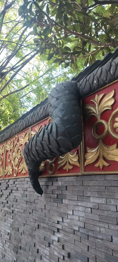 The image depicts the underside of a building structure in Tetela Del Monte, Mexico. The structure is adorned with a decorative frieze composed of repeating red panels with gold floral motifs, bordered by a dark gray, scaled texture. A curved, dark gray element that appears to be a stylized horn or tusk protrudes from the structure. Adjacent to the frieze, a wall constructed of gray brick or stone is visible. Trees and foliage appear in the background. The image is oriented at an angle, with the ground likely below the brick or stone wall. No people are visible in the frame.
