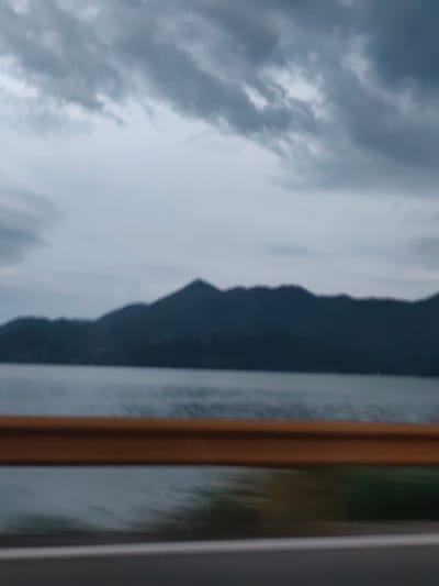 A wide shot captures a slightly blurred, overcast sky over a calm body of water with a dark, silhouetted mountain range in the distance. In the foreground, a bright orange guardrail is visible across the bottom third of the image, with a grey road surface below it. The scene appears to be located near Vranjina, Montenegro, as suggested by the mountainous landscape and body of water, likely Skadar Lake. No people or specific objects are discernible due to the blurriness and distance. The overall mood is somber, likely due to the cloudy weather.