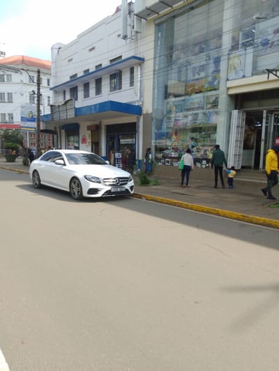 The image shows a white Mercedes-Benz car parked on the side of a paved road in Athi River, Kenya, at the coordinates -1.453, 37.007. The car is facing towards the right side of the image, with its front end visible. The road is empty except for the car. The road is bordered by a yellow line. The sidewalk is bordered by a concrete curb. On the right side of the image, there is a building with large storefront windows. The windows are filled with various items, including some toys, and are reflecting the sunlight. Two men and two women are standing in front of the store, looking at something off-camera. One man is wearing a black shirt and black pants, another man is wearing a black shirt and black shorts. One woman is wearing a black dress, and another woman is wearing a red shirt and blue jeans. There is a small child standing near one of the men, also looking off-camera. The child is wearing a black shirt and blue pants. The store is painted white with a blue awning. The building on the left side of the image is painted white with a blue awning. The sky is overcast. The scene is one of a typical sunny day in Athi River.
