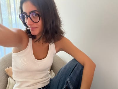 A female individual is depicted in an indoor setting located in Aldoar, Foz do Douro e Nevogilde, Portugal. The subject, positioned centrally, is seated and appears to be taking a selfie with her right arm extended, holding an unseen device. She has shoulder-length dark hair, a medium complexion, and is wearing round, tortoise-shell framed eyeglasses. Her attire consists of a white, ribbed scoop-neck tank top and dark blue denim jeans. A thin, gold-colored necklace is visible. She is seated in a light grey upholstered armchair, with a light beige decorative cushion positioned on its armrest or lap. Behind the subject, to the left, a window with sheer light-colored curtains is partially visible, indicating a source of natural light. The background to the right consists of a plain white wall. The subject's gaze is directed towards the camera.