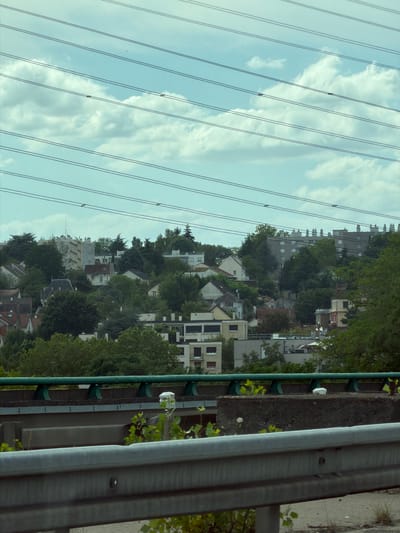 This media file depicts a daytime view of a hillside residential area in Palaiseau, France, seen from a slightly elevated perspective. The scene is dominated by several parallel power lines that traverse the frame diagonally from bottom left to top right, with gaps of blue sky and fluffy white clouds visible between them.

On the hillside, a dense cluster of buildings and trees creates a layered effect. The buildings range in size and architectural style, with some appearing as apartment blocks with many windows and balconies, while others are smaller, more traditional houses with pitched roofs. Lush green trees are interspersed throughout the residential area, suggesting a well-established, possibly suburban or semi-rural setting. The vegetation appears healthy and vibrant, indicating a likely warm season.

In the foreground, on the right side of the image, a metal guardrail is visible, suggesting the photo was taken from a road or elevated walkway. The guardrail is grey and appears to be made of corrugated metal. A dark green pipe runs parallel to the guardrail, supported by dark green brackets.

The overall impression is one of a tranquil, everyday scene in a French town, possibly during a sunny or partly cloudy day. There are no people or overt activities visible, lending a static and observational quality to the image. The light suggests it is daytime, with no specific time of day being discernible. No text is visible in the image.