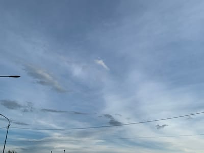 The image shows a cloudy sky over Pisochyn, Ukraine. The sky is a mix of pale blue and gray, with streaks of white clouds. There are two streetlights visible in the lower left portion of the image, and power lines stretch across the frame. The overall impression is of an overcast day.