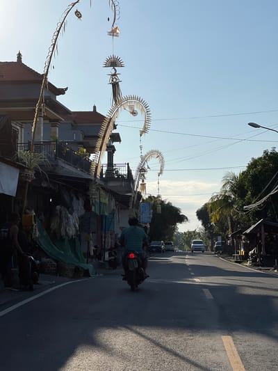 A street scene in Kubu, Indonesia, features traditional Balinese decorations arching over the road. These decorations, known as "penjor," are made of woven palm leaves and other natural materials, and are often erected during religious festivals. The scene is captured in the early morning light, casting long shadows across the asphalt. A lone motorcyclist is seen riding away from the camera, heading down the road. Several parked cars and a few buildings line the street, suggesting a small town or village setting. The sky is clear blue, with a few wispy clouds. The overall impression is one of a peaceful and vibrant community, with a strong connection to its cultural heritage.
