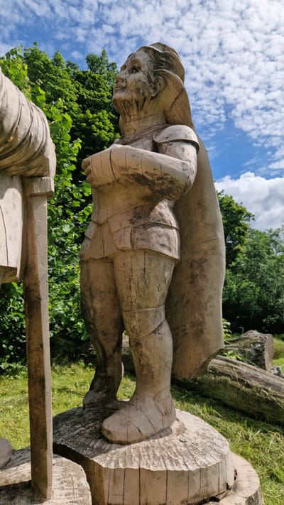 A weathered wooden sculpture of a person in armor stands outdoors on a wooden base. The sculpture is light brown and shows significant cracking and wear, suggesting exposure to the elements. The figure is depicted with its arms crossed and is wearing a helmet, shoulder armor, and segmented lower body armor. A cape is draped over its back. The sculpture is positioned in a grassy area with lush green trees in the background under a partly cloudy blue sky. Fallen logs are visible in the grass behind the sculpture. The sculpture is part of the sculpture park in Embūte, Latvia.