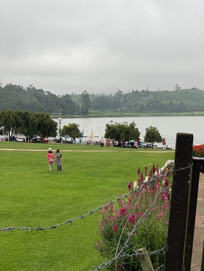 The image shows a landscape scene in Nuwara Eliya, Sri Lanka. In the foreground, there is a green lawn with two people on it. A barbed wire fence and wooden structure are partially visible on the right side of the frame. A line of trees separates the lawn from a body of water. Across the water, there are hills covered with trees and possibly tea plantations, fading into a misty, overcast sky. There are a few small structures and what appear to be docks along the water's edge. Some people are near the water. The scene conveys an impression of a cloudy, somewhat cool day.