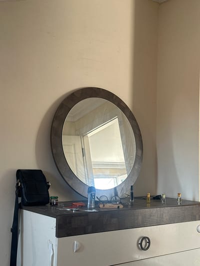 An indoor, medium shot captures a dresser with a round mirror above it. The dresser has a dark wood top and white drawers. On the dresser's surface, a black leather purse rests on the left. To its right are a small jar, some colorful wrappers, a pair of glasses on a wooden stand, a black bottle, and four small perfume bottles. A roll-on deodorant is also visible. The background is a plain, light-colored wall. The scene appears to be located in a bedroom or dressing area, likely within a residential building in Başakşehir, Turkey, given the provided context. There are no people or direct interactions depicted.