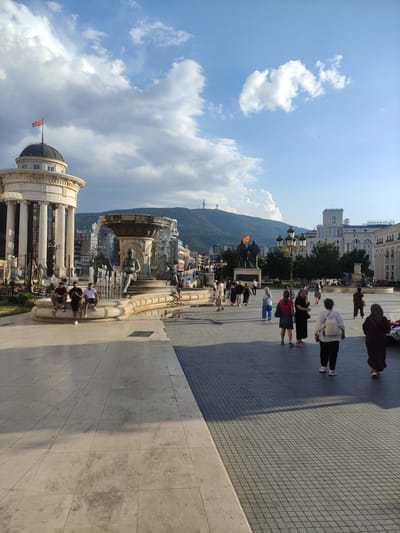 The image depicts Skopje, North Macedonia, specifically the Macedonia Square. In the foreground, large stone tiles form a spacious plaza. A textured, dark grey tiled pathway runs along the right side of the square. Several people are present, walking and sitting. Three men are seated on the edge of a large fountain in the mid-ground. The fountain itself is ornate, featuring sculptures and flowing water. To the left of the fountain stands a white building with classical columns and a dome topped with the flag of North Macedonia. In the background, buildings of various architectural styles are visible, along with a mountain range. A prominent cross statue is perched atop one of the hills. The sky is a mix of blue with scattered clouds, and the lighting suggests it is either early morning or late afternoon.